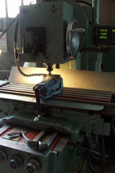 Frezarka pionowa z zamocowanym imadłem, obrabiająca metalowy element, widoczny panel sterowania z wyświetlanymi wartościami.