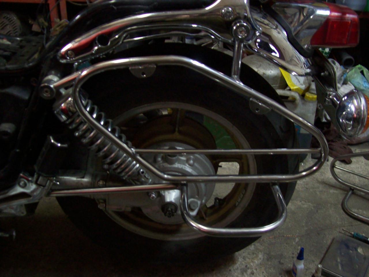 Chromowana konstrukcja rurowa, prawdopodobnie bagażnik, zamontowana na motocyklu, widoczny fragment tylnego koła i amortyzatora, otoczenie warsztatowe.