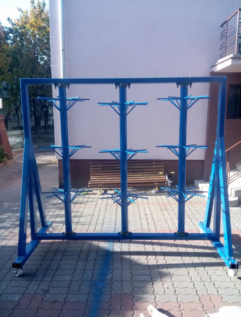 Niebieski, metalowy stojak na kółkach z trzema rzędami poziomych ramion, wykonany na zamówienie, ustawiony na brukowanym dziedzińcu przed budynkiem.