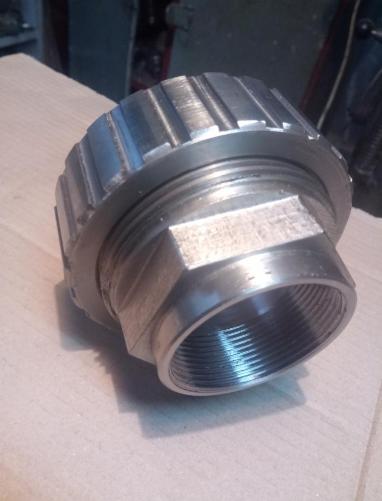 Metalowa nakrętka z gwintem wewnętrznym i sześciokątną podstawą, sfotografowana z bliska na kartonowym podłożu, widoczne ślady obróbki.