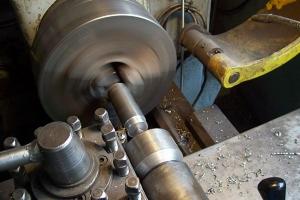 Tokarka w trakcie obróbki metalowego wałka, widoczne wióry i fragment żółtego elementu konstrukcyjnego maszyny.