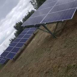 Rząd paneli słonecznych ustawionych na metalowych stelażach na trawiastym terenie, widoczne słupy wysokiego napięcia w tle pod pochmurnym niebem.