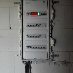 Rozdzielnica elektryczna zamontowana na ścianie z betonu komórkowego, uszczelniona pianką montażową, z widocznymi przewodami oznaczonymi numerami i zielonymi diodami.