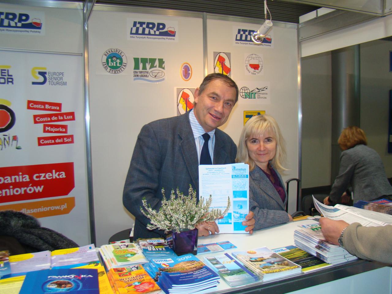 Stoisko targowe z ulotkami i folderami o tematyce turystycznej, prezentacja oferty wyjazdów dla seniorów, mężczyzna w garniturze i kobieta prezentują folder z ofertą Sylwestra w Czechach, w tle...