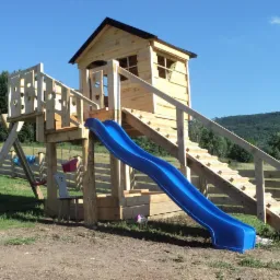 Drewniany plac zabaw z domkiem, zjeżdżalnią i huśtawkami na tle zielonego wzgórza.