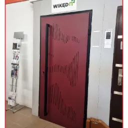 Wystawowa, bordowa, antywłamaniowa drzwi z długim, pionowym uchwytem i wyciętym wzorem z pionowych kresek, prezentowana w salonie obok stojaka z ulotkami i fragmentu innej ekspozycji drzwi.
