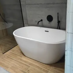 Nowoczesna, wolnostojąca wanna z czarną armaturą na tle betonowej ściany i drewnopodobnych płytek podłogowych, z widocznym fragmentem szklanej kabiny prysznicowej.