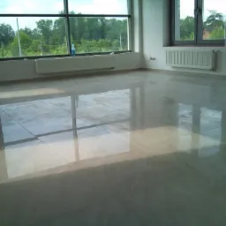 Błyszcząca, beżowa podłoga z płytek w nowym, pustym pomieszczeniu; duże okna z widokiem na zieleń, białe grzejniki pod oknami; odbicia światła na podłodze.