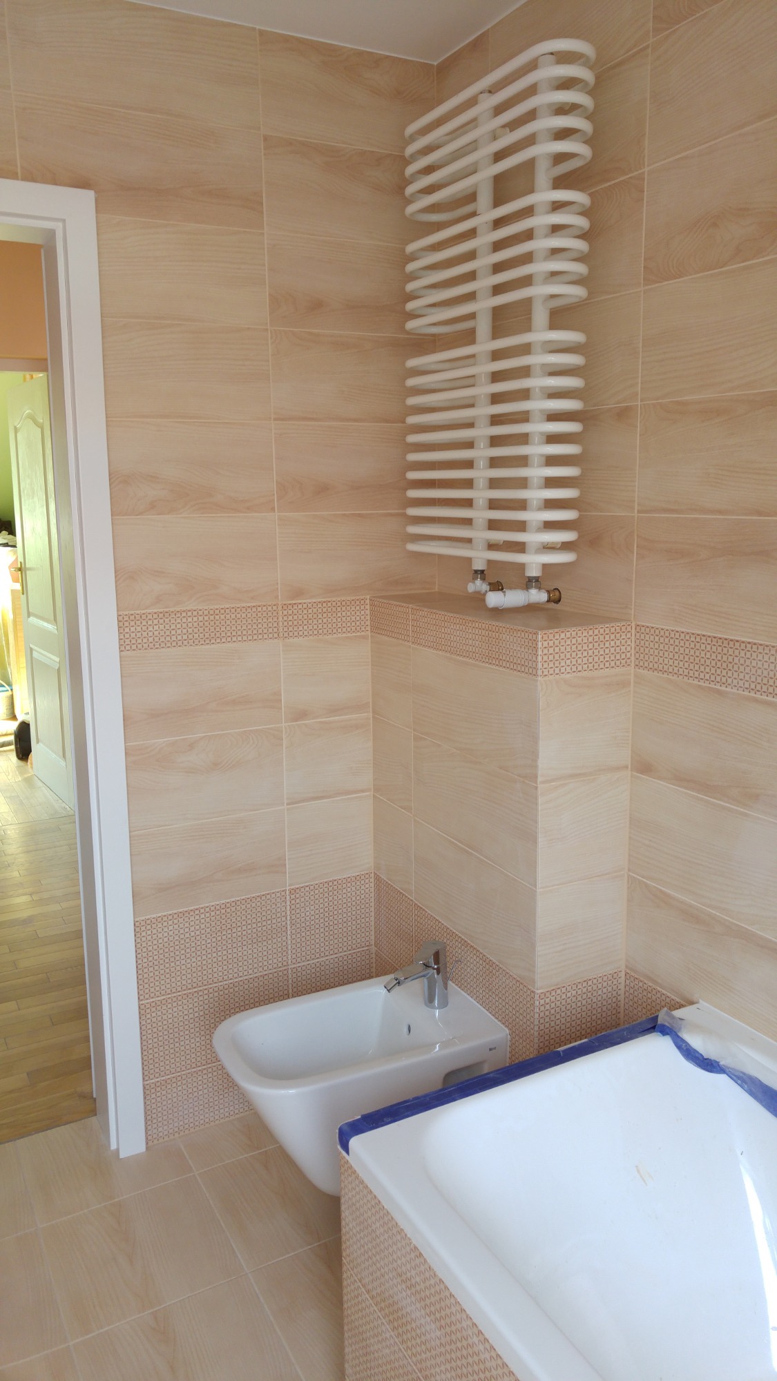Jasna łazienka z beżowymi płytkami imitującymi drewno, spiralny grzejnik drabinkowy, białe bidet i wanna zabezpieczona folią malarską.