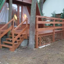 Drewniany taras z ażurową balustradą i schodami przy domku letniskowym, otoczony drzewami.