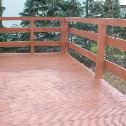 Drewniany taras z brązową balustradą i deskami podłogowymi, widok na zieleń drzew w tle.