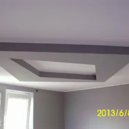 Szara, podwieszana konstrukcja sufitowa w kształcie kwadratu z otworem, widoczna z dołu. W tle okno i ściana w odcieniach szarości. Data w prawym dolnym rogu.