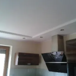 Gładki, biały sufit z oświetleniem punktowym, wkomponowany w nowoczesną kuchnię z ciemnymi szafkami i okapem. Widoczne detale wykończenia.