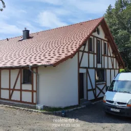 Budynek z elewacją w stylu szachulcowym, świeżo po malowaniu, z zaparkowanym białym vanem i pracującym mężczyzną przy klimatyzatorze na zewnątrz.