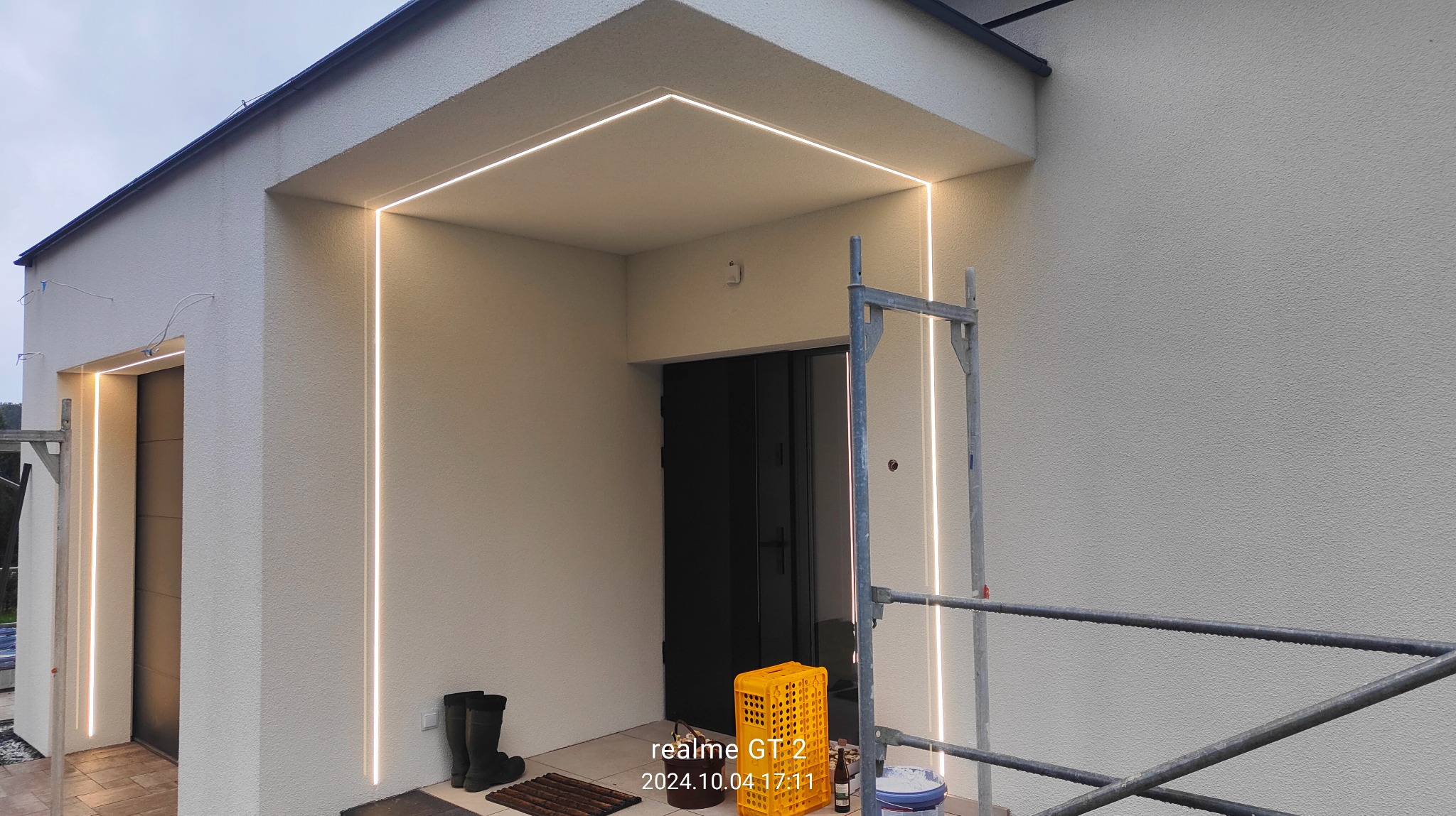 Nowoczesna elewacja domu z oświetleniem LED w kształcie ramy wokół wejścia, widoczne rusztowanie, gumowce i wiadro z farbą sugerujące prace wykończeniowe.