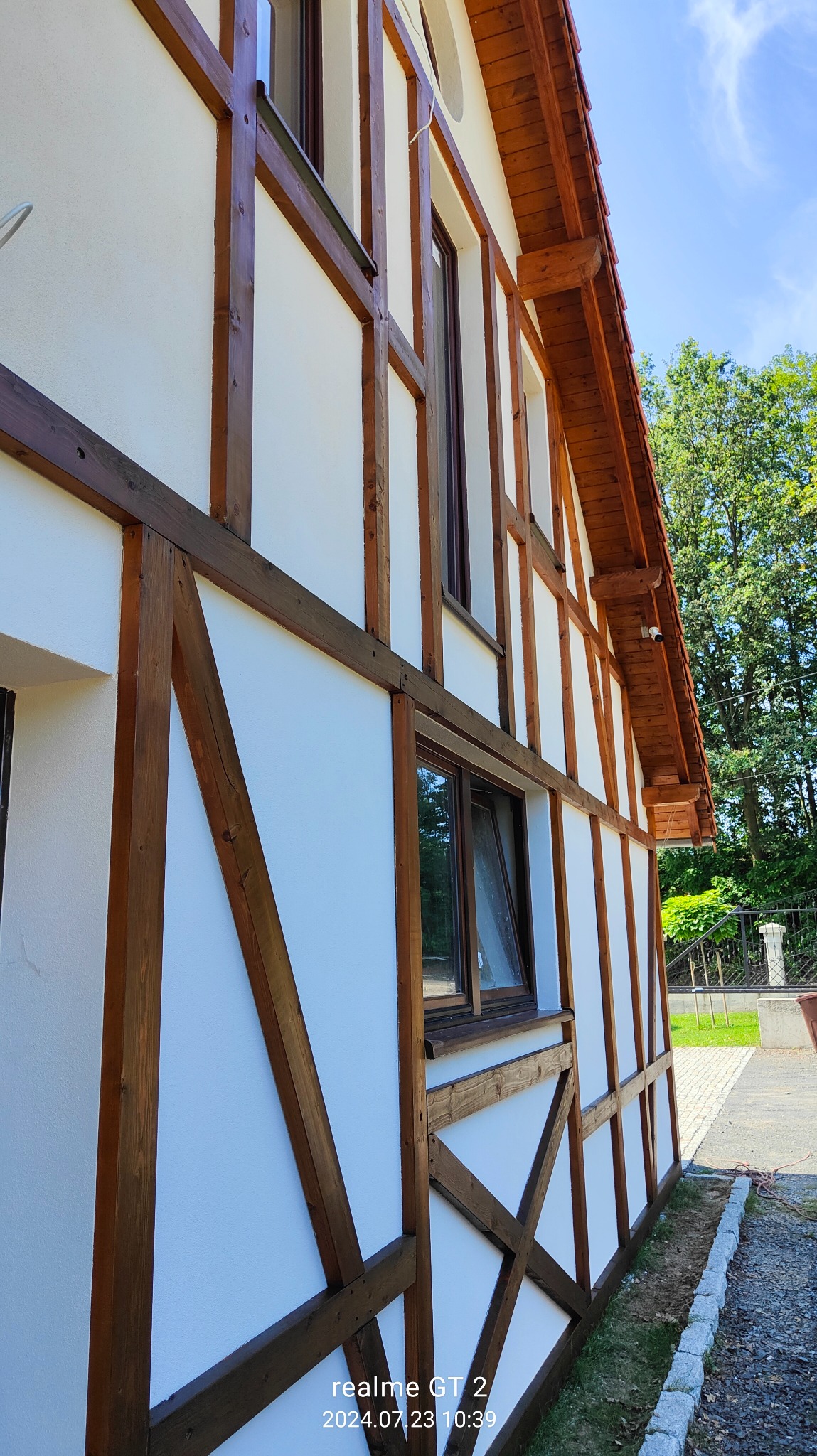 Boczny widok domu z białą elewacją w stylu szachulcowym z ciemnobrązowymi drewnianymi belkami, widoczne okna i fragment dachu z brązową dachówką, w tle zieleń drzew i fragment chodnika.