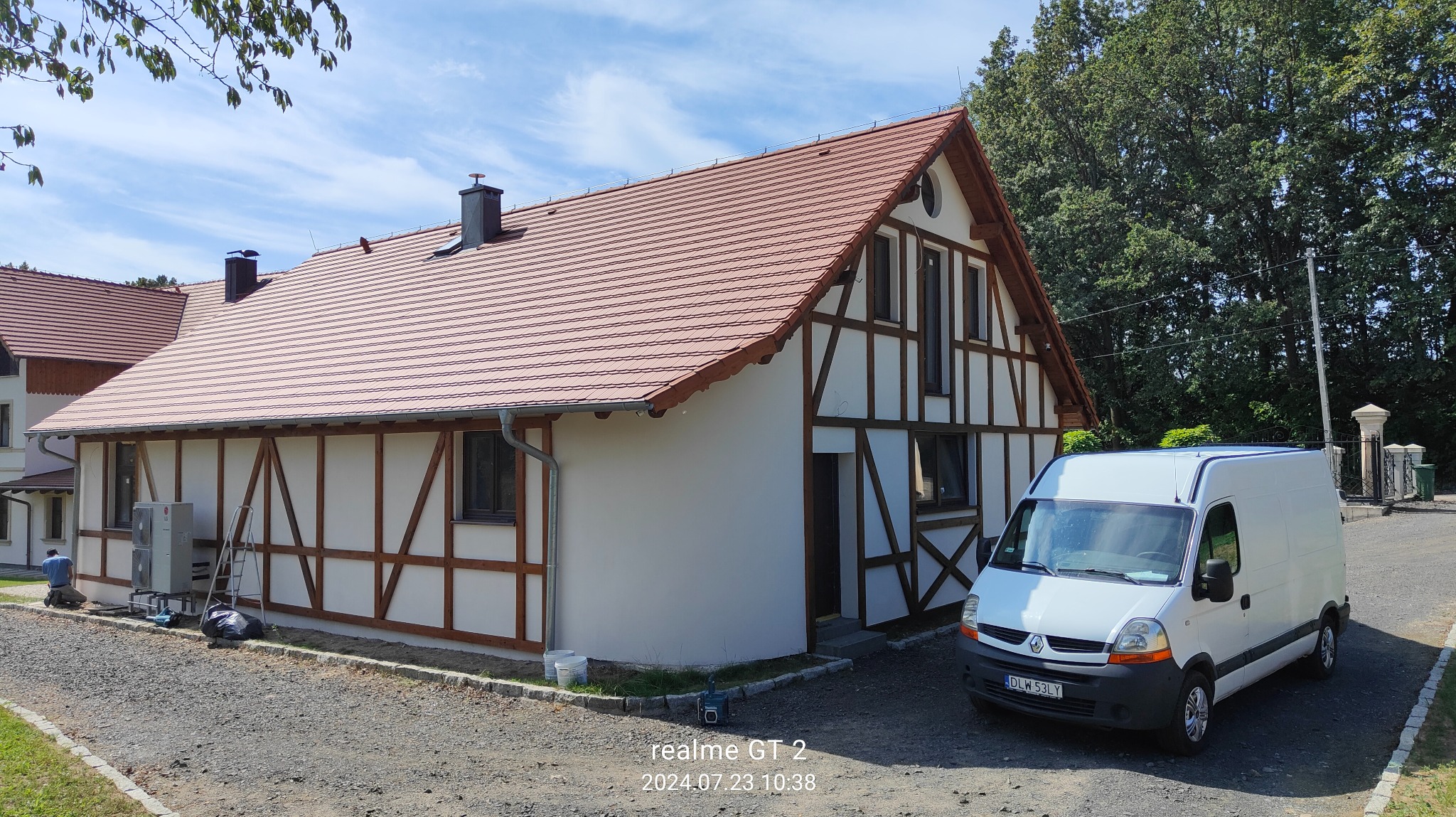 Budynek z elewacją w stylu szachulcowym, świeżo po malowaniu, z zaparkowanym białym vanem i pracującym mężczyzną przy klimatyzatorze na zewnątrz.