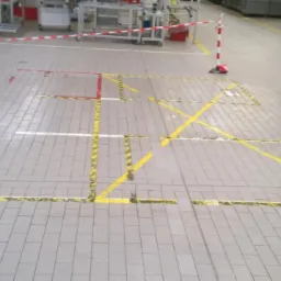 Szara, przemysłowa posadzka z kostki, z widocznymi żółto-czarnymi pasami taśmy ostrzegawczej wyznaczającej obszary i linie. Fragmenty starego, czerwonego i białego oznakowania miejscami widoczne.