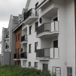 Nowoczesny budynek wielorodzinny z balkonami zabezpieczonymi czarnymi, metalowymi balustradami, widok z poziomu gruntu na tle pochmurnego nieba.