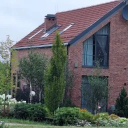 Dwukondygnacyjny dom z czerwonej cegły z oknami dachowymi i dużym oknem w szczycie, otoczony zielenią. Widoczny komin i antena satelitarna.