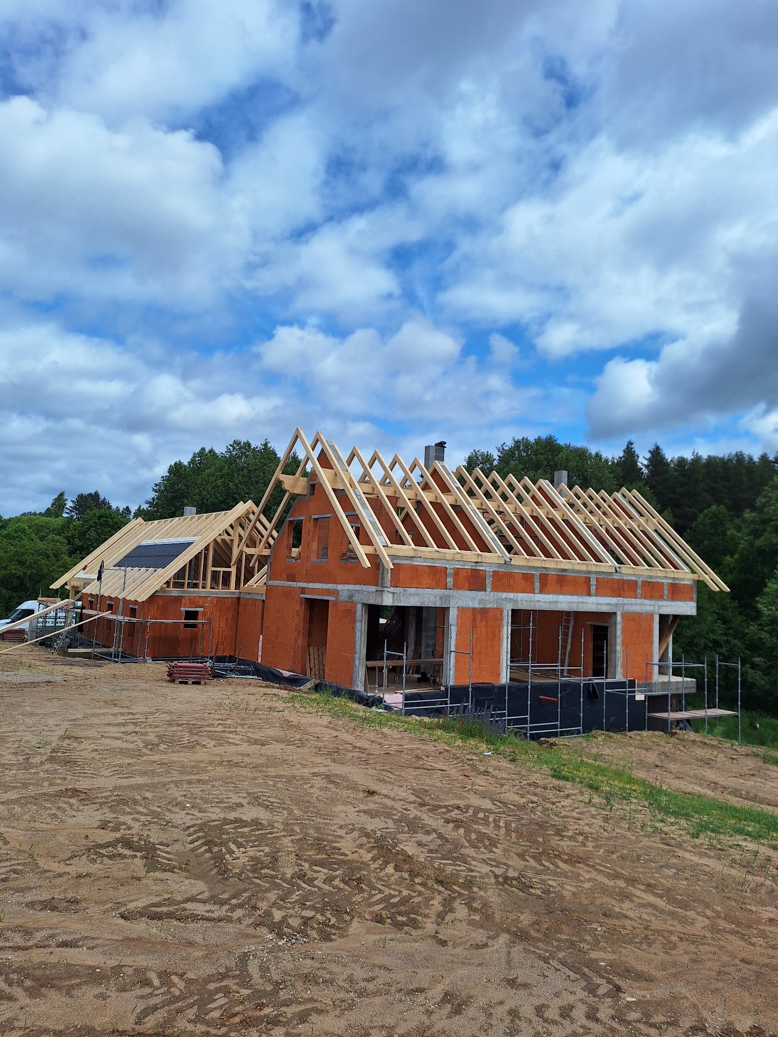Budowa domu z czerwonej cegły w trakcie wznoszenia dachu, widok z dołu na tle błękitnego nieba z chmurami, teren budowy z ziemią.