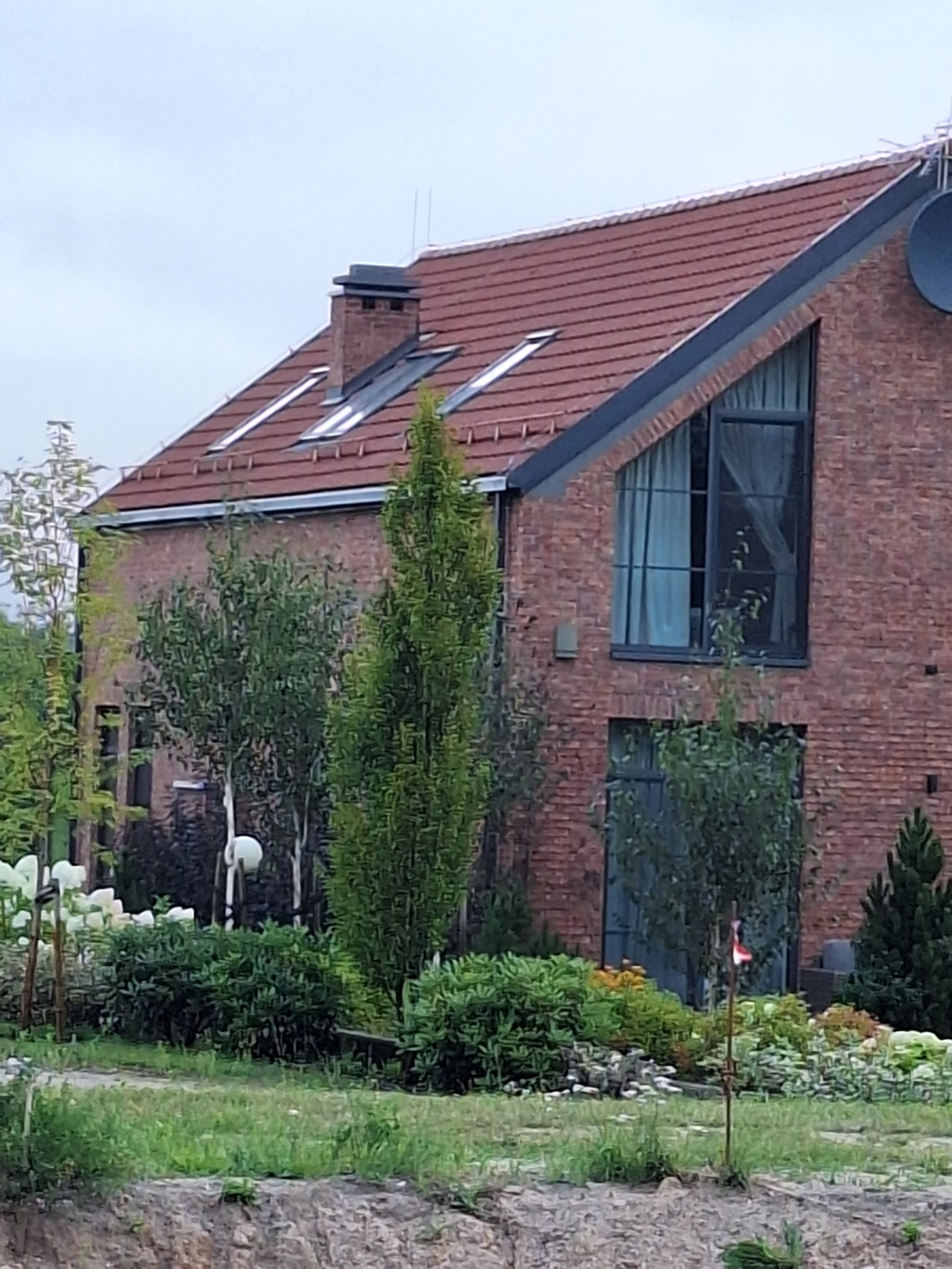 Dwukondygnacyjny dom z czerwonej cegły z oknami dachowymi i dużym oknem w szczycie, otoczony zielenią. Widoczny komin i antena satelitarna.