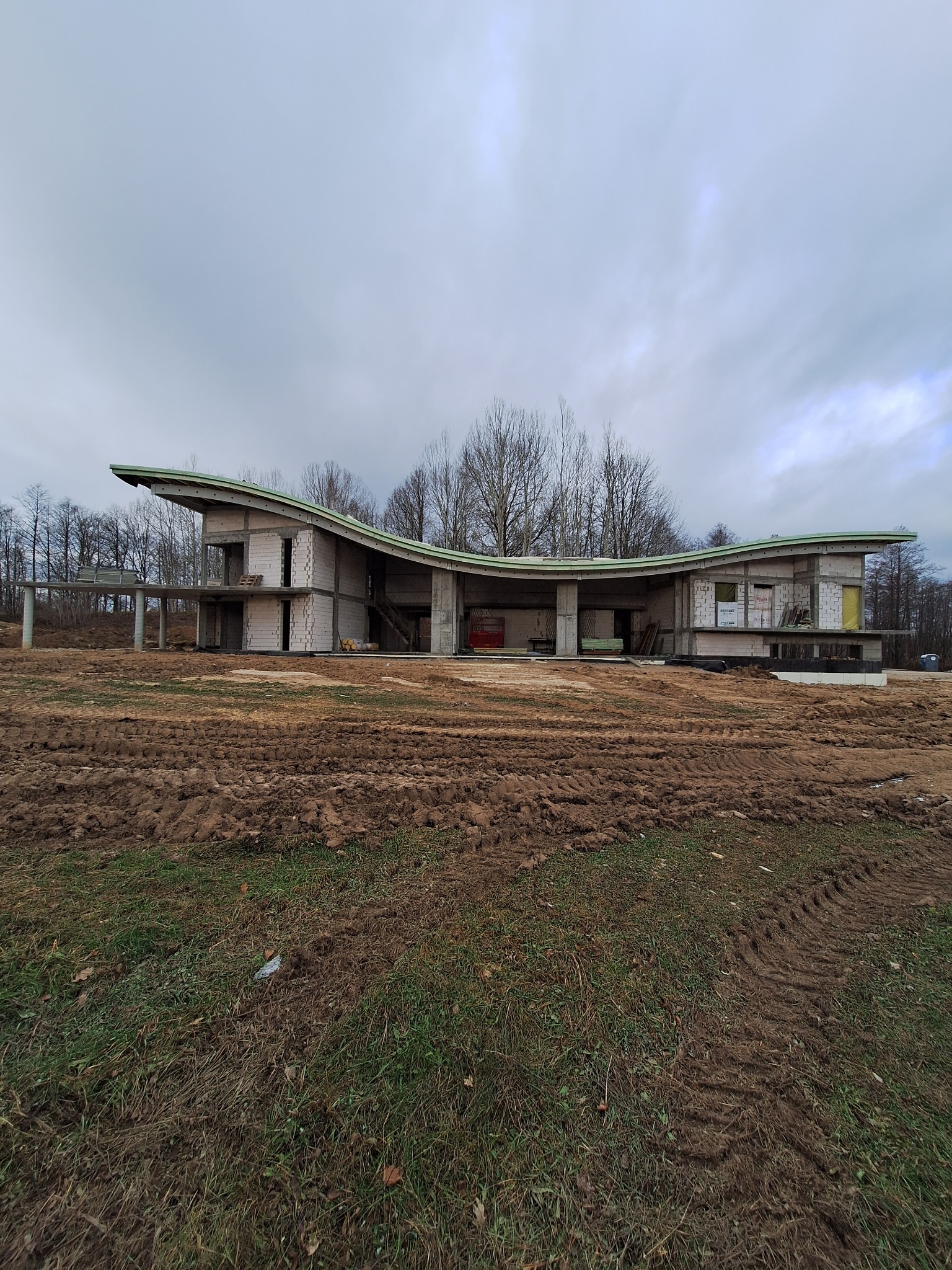 Niedokończony, parterowy dom z betonu komórkowego o falistym dachu, widoczny na tle drzew i pochmurnego nieba. Teren wokół budynku jest nierówny, pokryty błotem.
