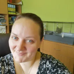 Uśmiechnięta kobieta z niebieskimi oczami w biurze, za nią regał z segregatorami i klatka dla zwierząt, co sugeruje przyjazne środowisko pracy.