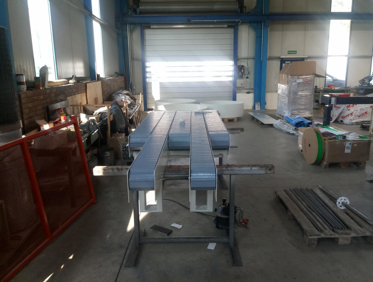 Wnętrze hali produkcyjnej z trzema równoległymi, metalowymi przenośnikami taśmowymi na stole montażowym, przygotowanymi do transportu wewnątrz zakładu.