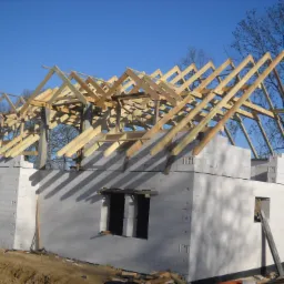 Wznoszenie konstrukcji dachu z drewnianych belek na surowym budynku z białych bloczków, widoczne otwory okienne i drzwiowe, w tle błękitne niebo.