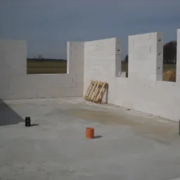 Wnętrze domu w trakcie budowy z białych bloczków, widoczne otwory okienne z widokiem na pole, na podłodze stoi mały głośnik i pomarańczowa rura wentylacyjna, oparta o ścianę drewniana konstrukcja.