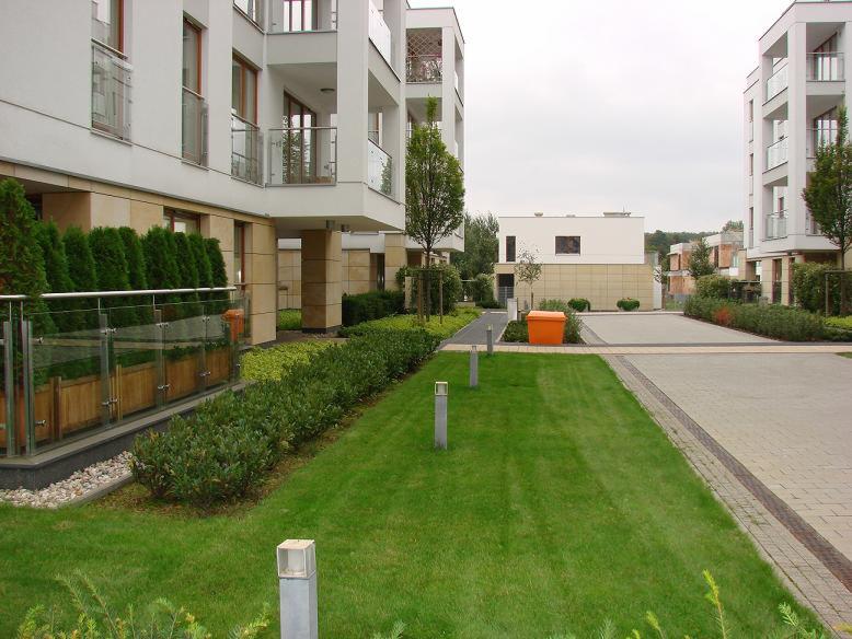 Nowoczesny kompleks apartamentowy z zadbanym trawnikiem, niskimi lampami oświetleniowymi i roślinnością ozdobną, pomarańczowy kontener na śmieci w tle.