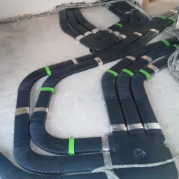 Czarny system kanałów wentylacyjnych z elementami mocującymi w kolorze srebrnym i zielonym, ułożony na surowej podłodze w trakcie montażu.