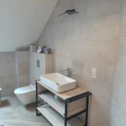 Nowoczesna łazienka na poddaszu z umywalką nablatową na drewnianym regale z czarnymi elementami, szare płytki na ścianach i podłodze imitujące drewno, toaleta podwieszana.