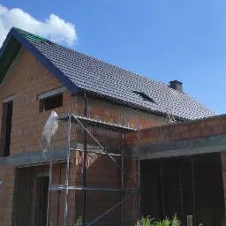 Wznoszenie ścian z czerwonej cegły na budowie domu jednorodzinnego, widoczne rusztowanie i częściowo położony dach z ciemnej dachówki na tle błękitnego nieba z chmurami.