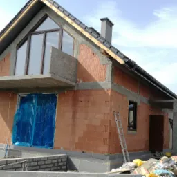 Budowa domu jednorodzinnego z czerwonej cegły ceramicznej; widoczne okna, balkon i zarys tarasu; pracownik budowlany miesza zaprawę w tle.