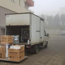 Rojson Trans - Ciężarówka z otwartą przestrzenią ładunkową na rampie, załadowana kartonami z nowymi kuchenkami mikrofalowymi i elektrycznymi marki Samsung, na tle zamglonego osiedla.