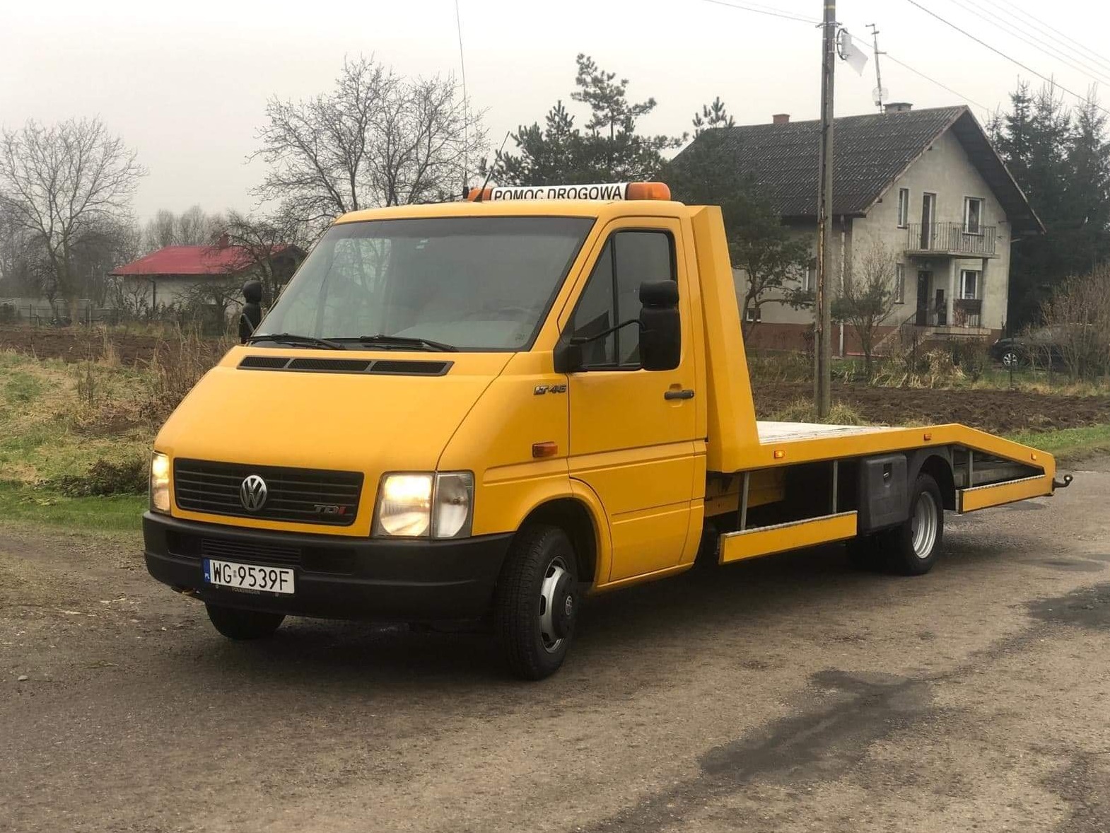 Żółta laweta Volkswagen LT 46 z napisem 'Pomoc Drogowa' na dachu, zaparkowana na szutrowej drodze, z widocznym numerem rejestracyjnym WG 9539F i domami w tle.