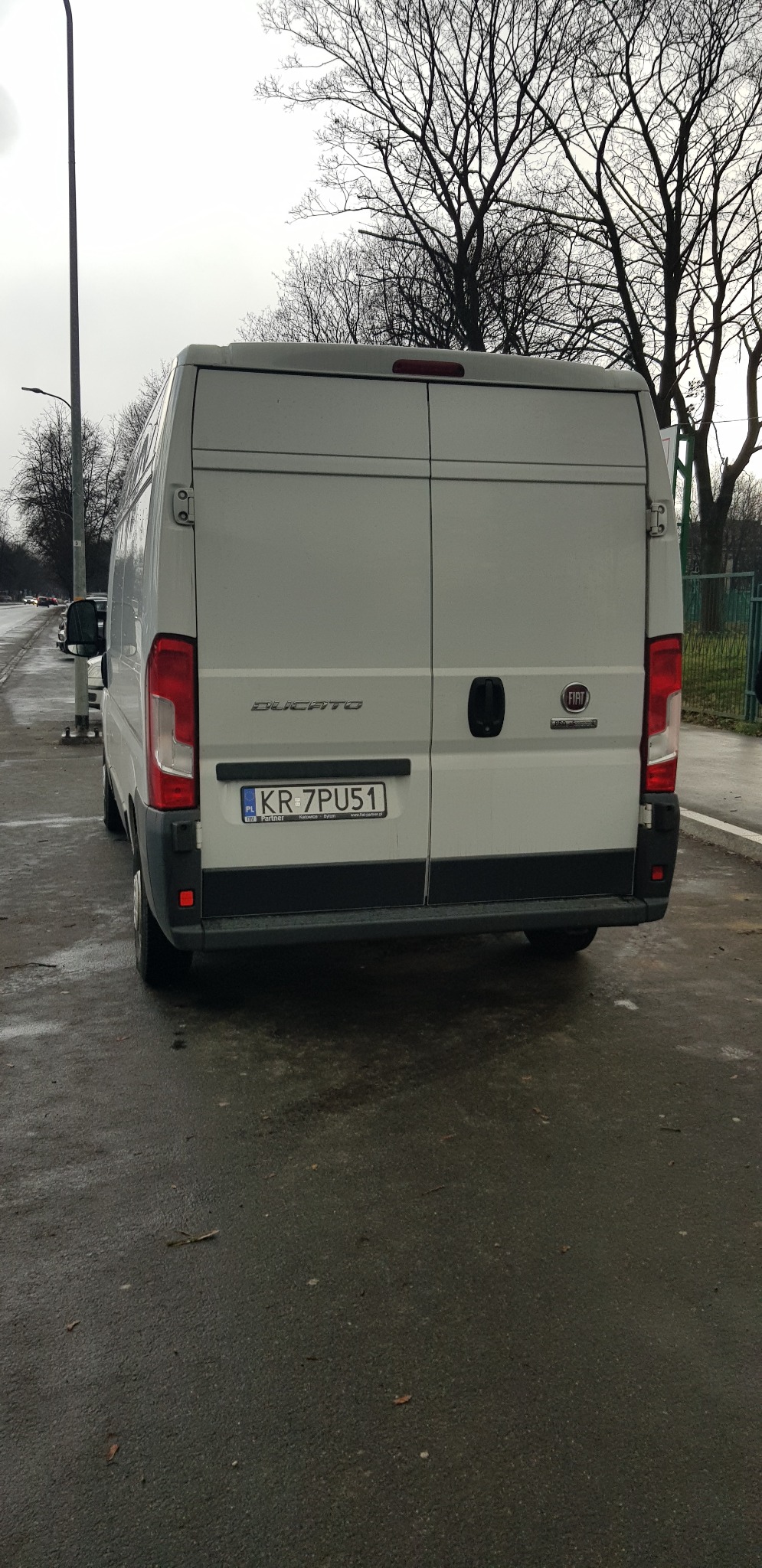 Tył białego furgonetki Fiat Ducato z polską tablicą rejestracyjną KR 7PU51, zaparkowanego na mokrym asfalcie w pochmurny dzień. W tle widoczne nagie drzewa.