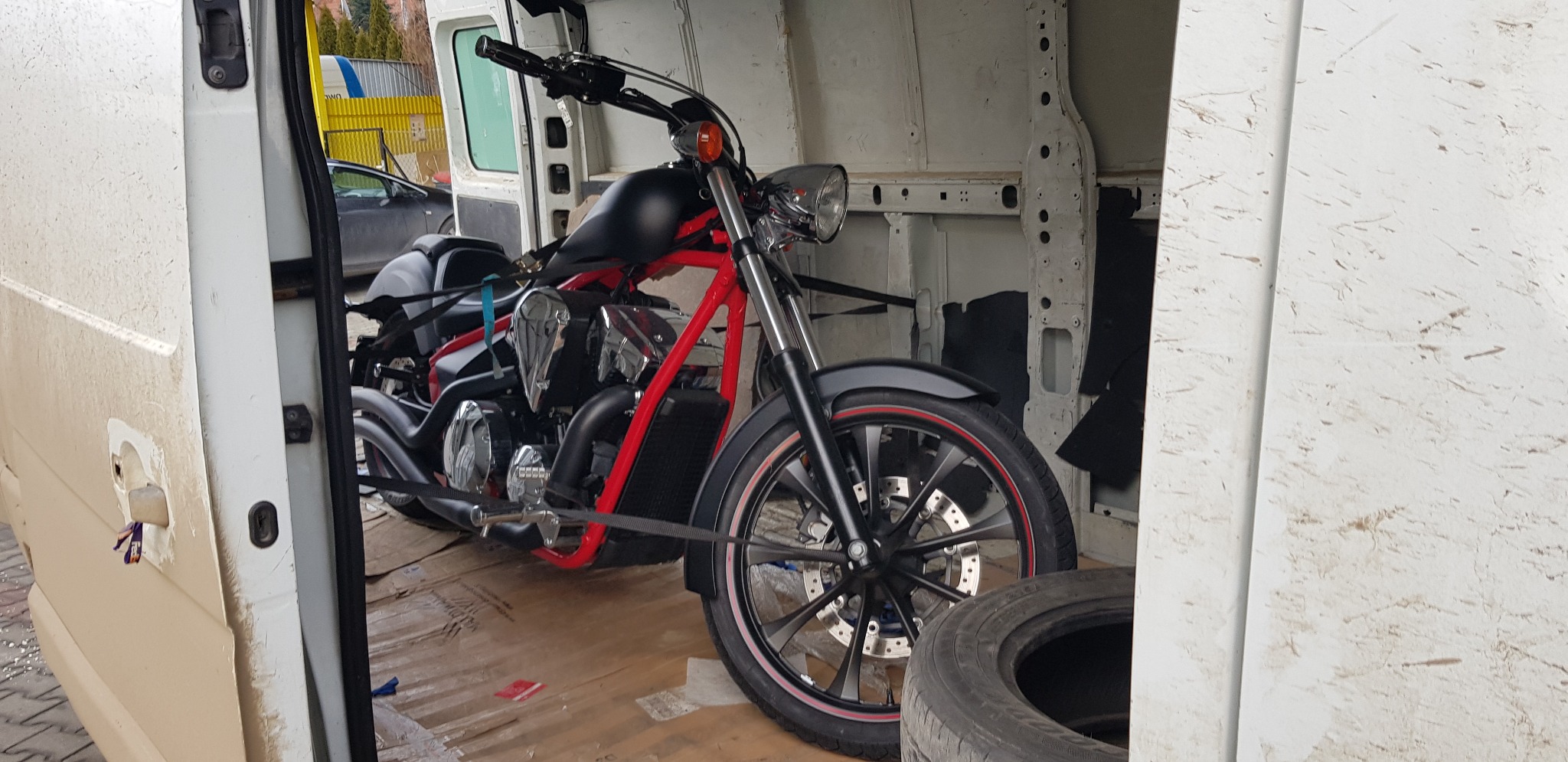 Czarny motocykl z czerwonymi elementami ramy, zabezpieczony pasami wewnątrz brudnego, białego furgonu. Widoczne opony obok motocykla.