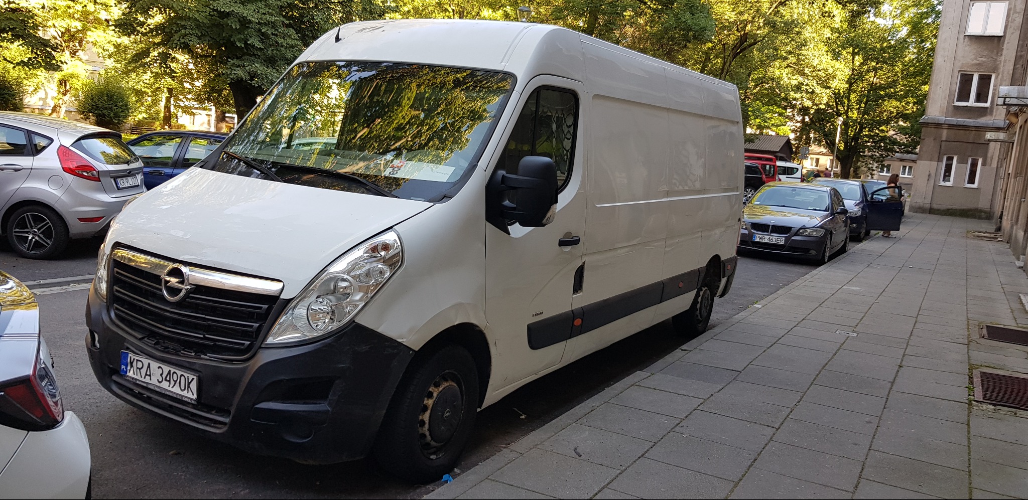 Biały van Opel Master zaparkowany na chodniku w mieście, z widocznym numerem rejestracyjnym KRA 3490K, odbicia drzew w szybie i inne samochody w tle.