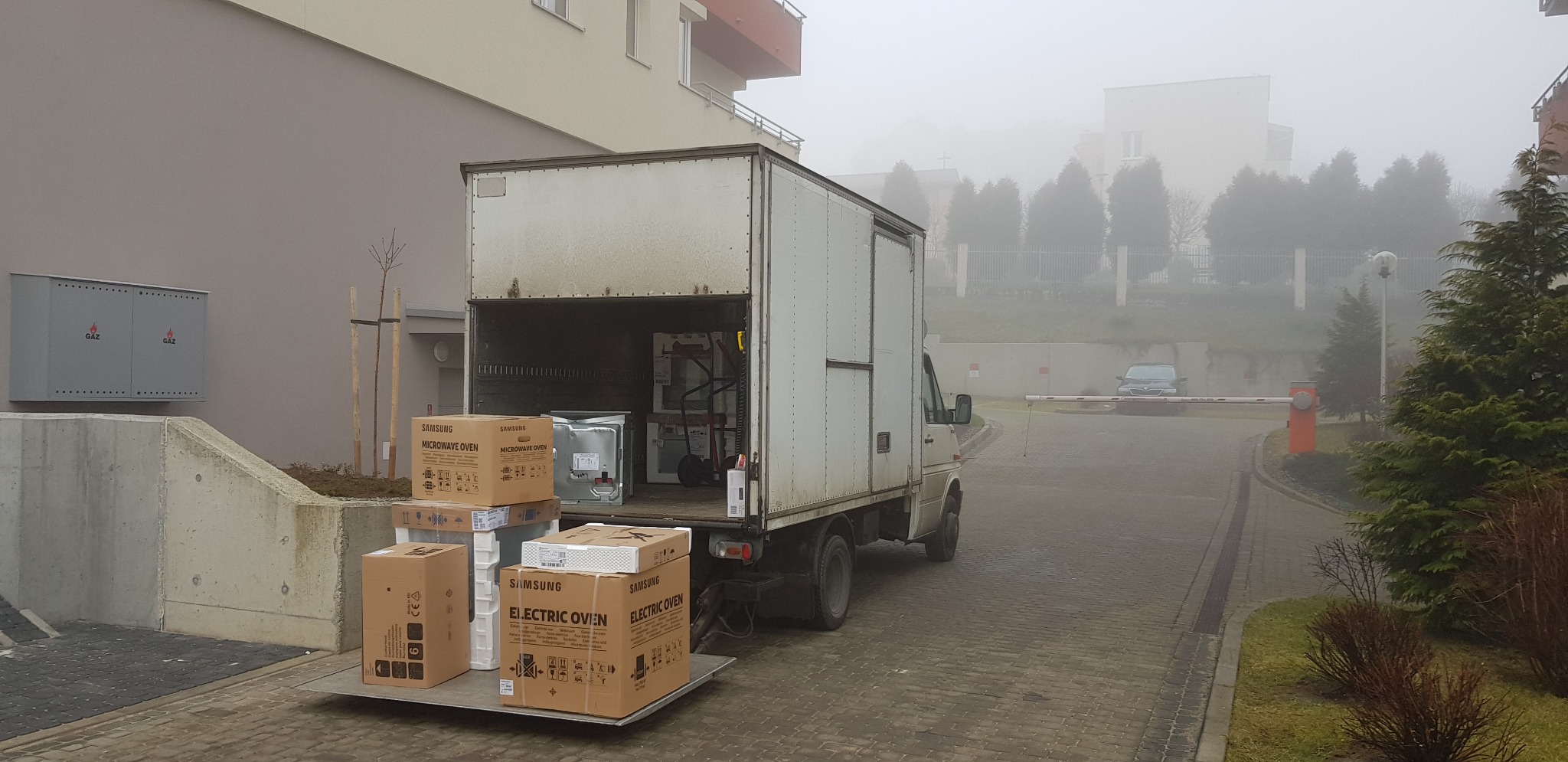 Ciężarówka z otwartą przestrzenią ładunkową na rampie, załadowana kartonami z nowymi kuchenkami mikrofalowymi i elektrycznymi marki Samsung, na tle zamglonego osiedla.