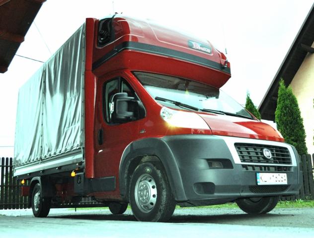 Czerwony samochód dostawczy Fiat Ducato z plandeką, zaparkowany na tle domu z tujami, widoczne tablice rejestracyjne.