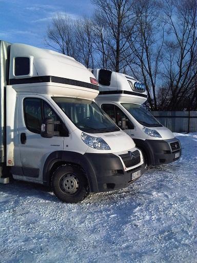 Dwa białe samochody dostawcze z zabudową typu furgon stojące na śniegu, widoczne z przodu pod kątem. Jeden z pojazdów ma dodatkowe oświetlenie na dachu.