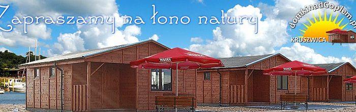 Rząd drewnianych domków letniskowych z czerwonymi parasolami Warka na tle błękitnego nieba z chmurami i częściowo widocznym jeziorem.