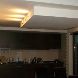 Nowoczesna kuchnia z podwieszonym sufitem i oświetleniem punktowym LED, widok na zabudowę meblową w ciemnym brązie, jasna ściana z cegiełkami i okno z żaluzjami.