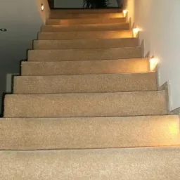 Schody wyłożone beżową wykładziną dywanową z oświetleniem punktowym wbudowanym w ścianę, dodatkowo poręcz ze stali nierdzewnej po prawej stronie.