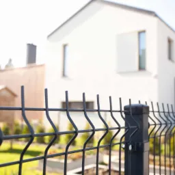 Ciemnoszare ogrodzenie panelowe na tle nowoczesnego, białego domu z ogrodem, widoczne z bliskiej perspektywy.