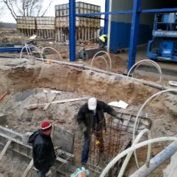 Dwóch robotników przy zbrojeniu fundamentów pod budynek przemysłowy, widoczne szalunki, wykop, rury, w tle konstrukcja stalowa i podnośnik koszowy.