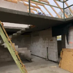 Wnętrze domu w trakcie budowy: surowe ściany z betonu kom&oacute;rkowego, betonowe schody w stanie surowym, drewniana konstrukcja dachu i balkonu, widoczna drabina oparta o schody, łopata oparta o ścianę.